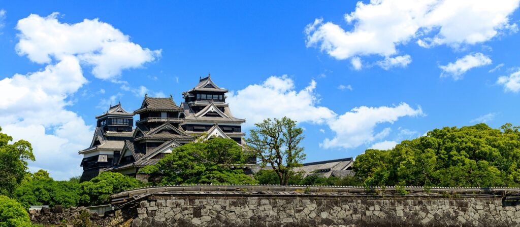 A traditional Japanese castle with dark roofs and multiple storeys stands surrounded by green trees and stone walls, set against a bright blue sky with scattered white clouds.