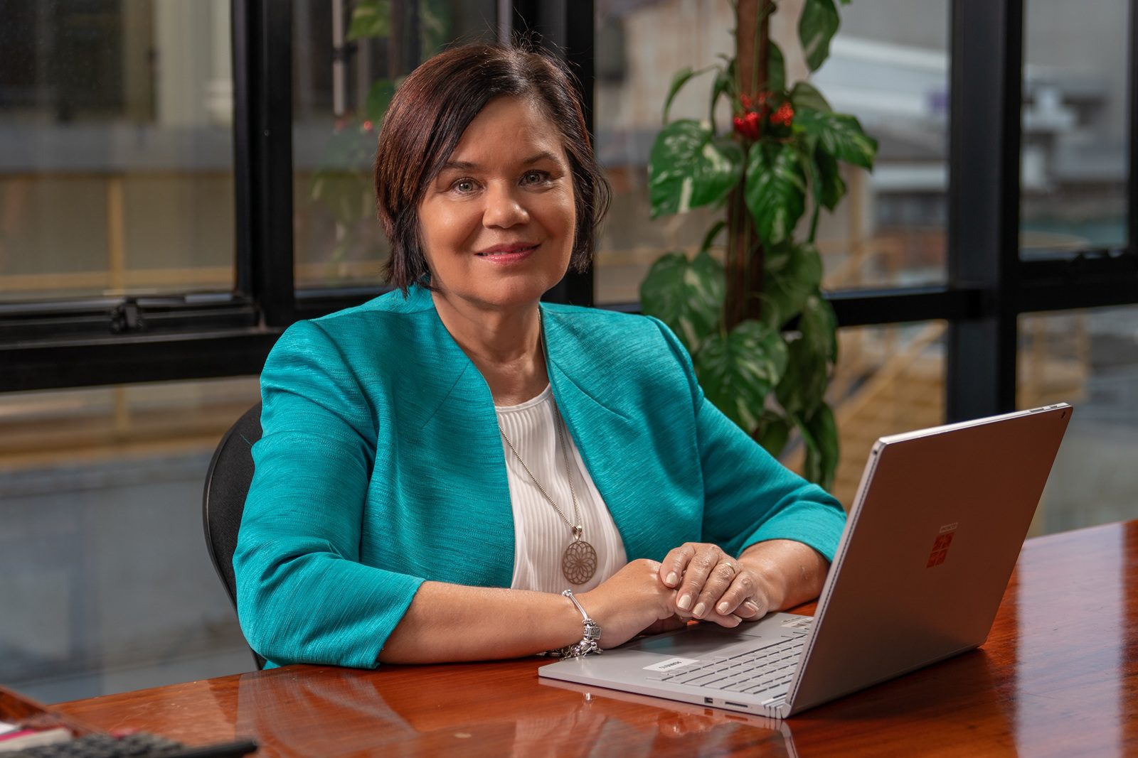 A woman with short brown hair, wearing a teal jacket and white blouse, sits at a wooden desk with a laptop. She is smiling and has her hands resting on the laptop. There’s a plant and large windows in the background.