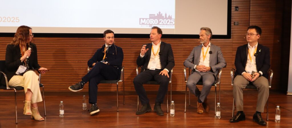 Five people sit on a stage in a panel discussion, four men and one woman, each holding a microphone. Bottles of water are on the floor, and a screen behind them displays MORO 2025.