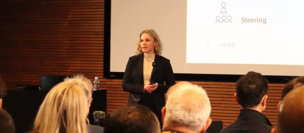 A woman stands in front of a seated audience giving a presentation. She is dressed in business attire, and a projection screen with text and graphics is visible behind her.