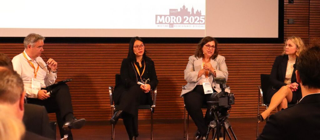 Four people sit on a stage in a panel discussion. Three women and one man are facing the audience, with the woman in grey speaking while others listen. A camera is positioned in front of them. The backdrop displays MORO 2025.