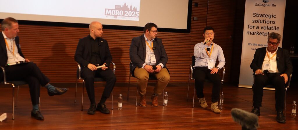 Five men sit on stage in a panel discussion. One man is speaking into a microphone. Behind them is a screen displaying “MORO 2025” and a banner that reads “Strategic solutions for a volatile marketplace”.