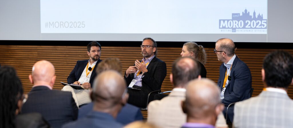 Four panellists sit on stage in front of an audience, engaged in discussion at a MORO 2025 event. One man speaks whilst the others listen. A screen behind them displays the event name and hashtag #MORO2025.