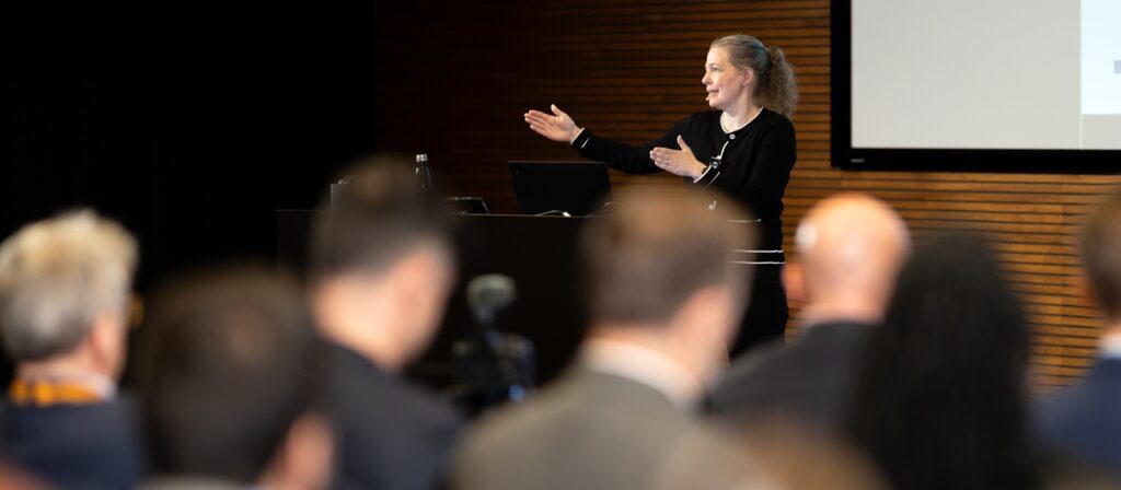 A woman stands at a lectern, gesturing with her hands whilst speaking to an audience. The crowd is seated and facing her, with a blurred focus on them and a clear view of the presenter.