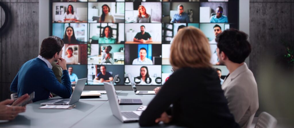 Four people sit at a conference table with laptops, participating in a virtual meeting displayed on a large screen showing many people in a video call grid.