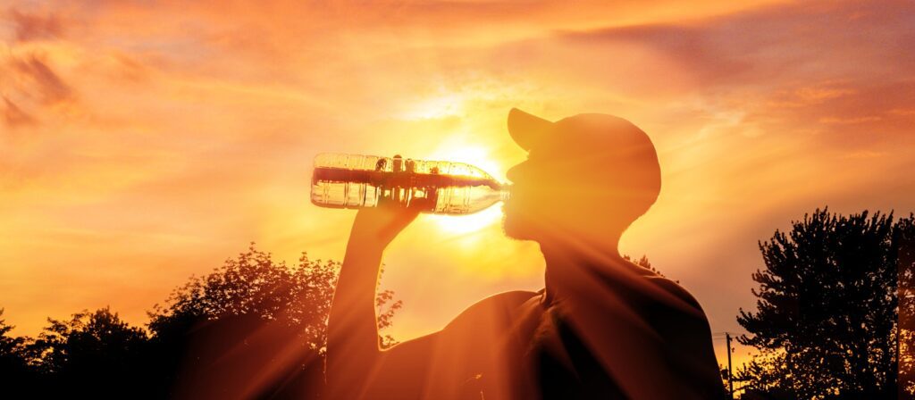 A person wearing a cap drinks from a water bottle outdoors at sunset, with the sun shining brightly behind them and trees silhouetted in the background.