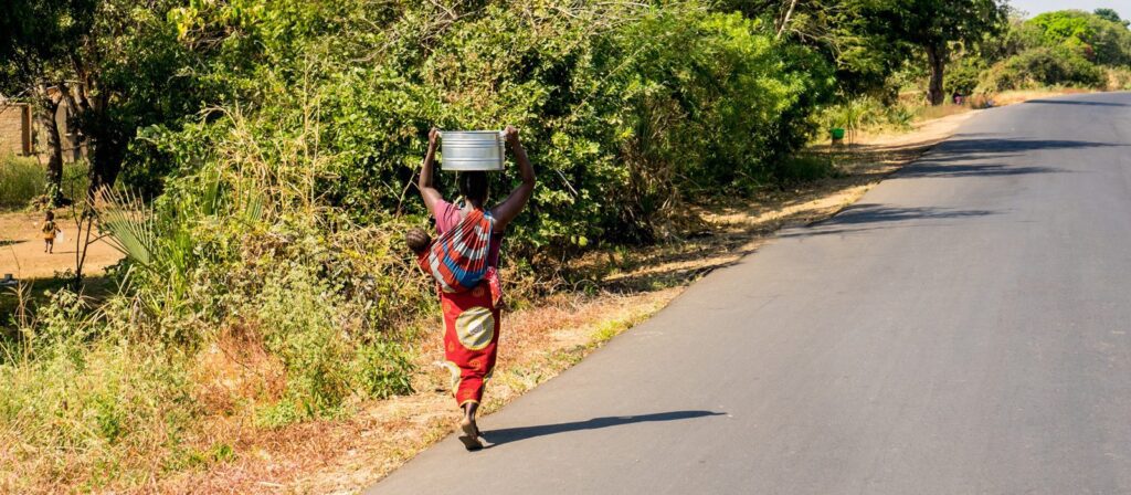 A person walks along a rural road carrying a metal basin on their head and a child on their back, surrounded by green vegetation and dry grass.