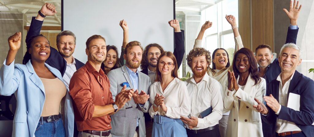 A diverse group of professionally dressed people stand together indoors, smiling, applauding, and raising their arms in celebration, suggesting a successful team achievement or event.