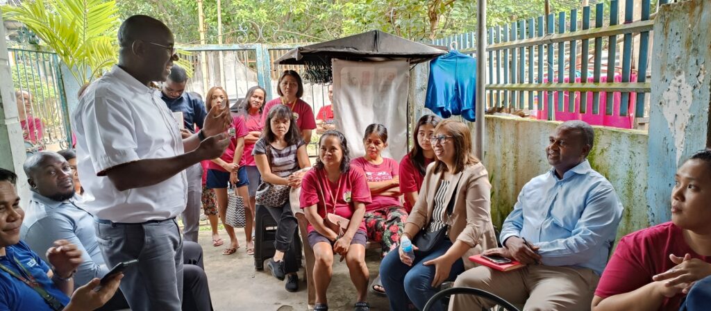 A man stands and speaks to a seated group of people, both men and women, in an outdoor setting with a fence and greenery in the background. Some people listen attentively whilst others hold notebooks or mobile phones.