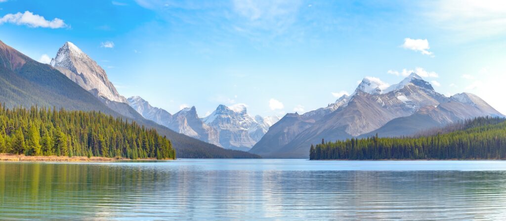 A panoramic view of a calm lake surrounded by dense evergreen forests, with snow-capped mountains rising in the background under a clear blue sky.
