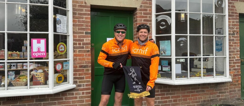 Two cyclists wearing matching orange and black jerseys and helmets stand smiling in front of a brick building with large windows and a green door, holding a dark-coloured banner with a white figure on it.