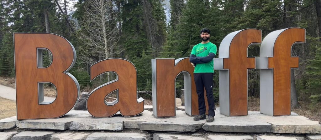 A person stands with arms crossed in front of a large Banff sign made of wood and metal, surrounded by trees and nature. The person is wearing a green shirt and dark trousers, and the scene appears to be outdoors.
