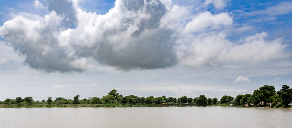 A calm lake with trees along the distant shore under a bright sky filled with large, fluffy clouds.