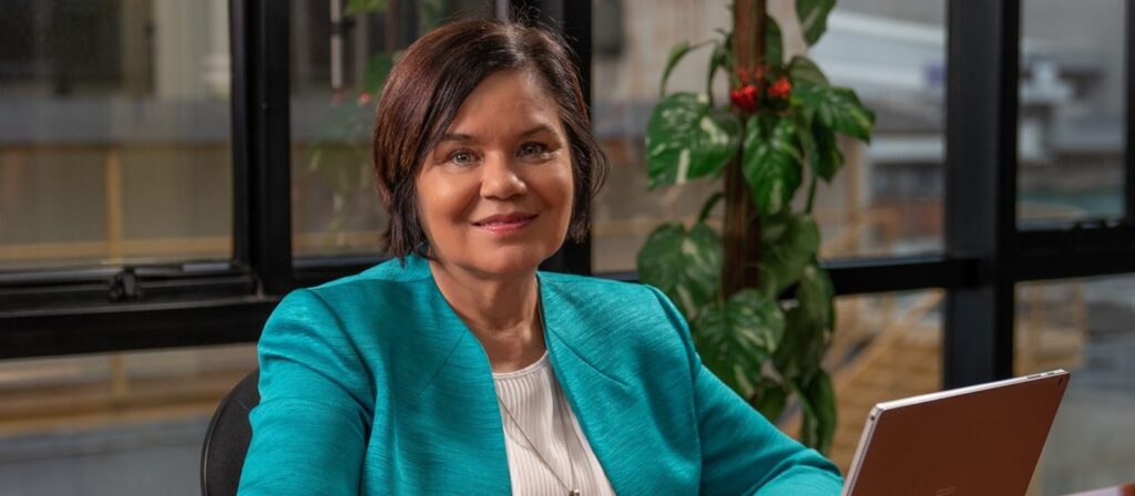 A woman with short brown hair in a teal blazer sits at a desk with a laptop, smiling at the camera. There is a plant and large windows in the background.