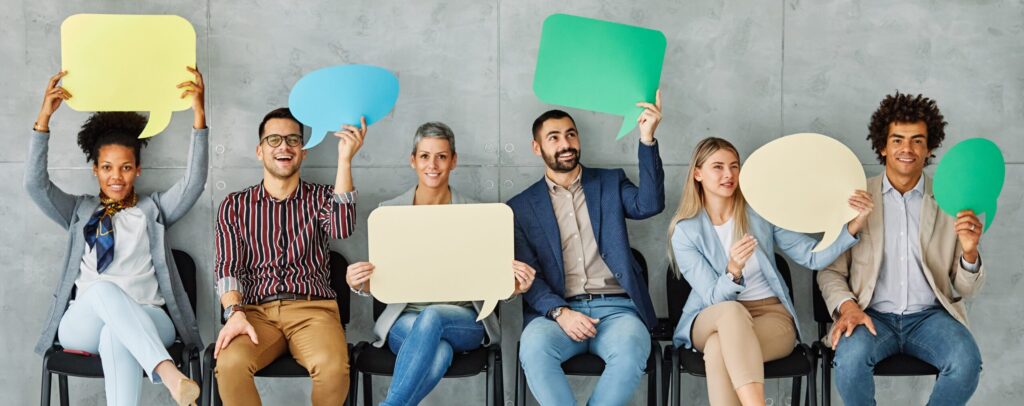 Six people sitting in a row holding colourful blank speech bubbles, smiling and looking at the camera, against a grey concrete wall background.