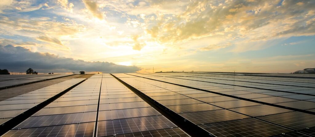 Rows of solar panels on a flat surface reflecting the sunlight, with a dramatic sky filled with clouds and the sun shining brightly near the horizon.