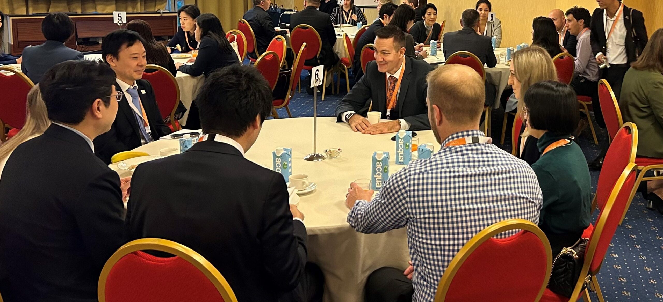 People in business attire sit around round tables having discussions at a conference or networking event. Bottled water, coffee cups, and notepads are on the tables. Red chairs and yellow walls are visible in the background.