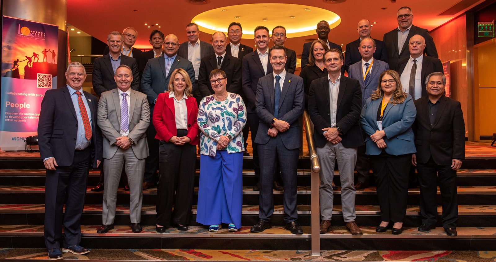 A group of about 22 people in business attire stand together on stairs indoors, smiling for a group photo at a professional event. A colourful event banner is visible on the left side of the image.