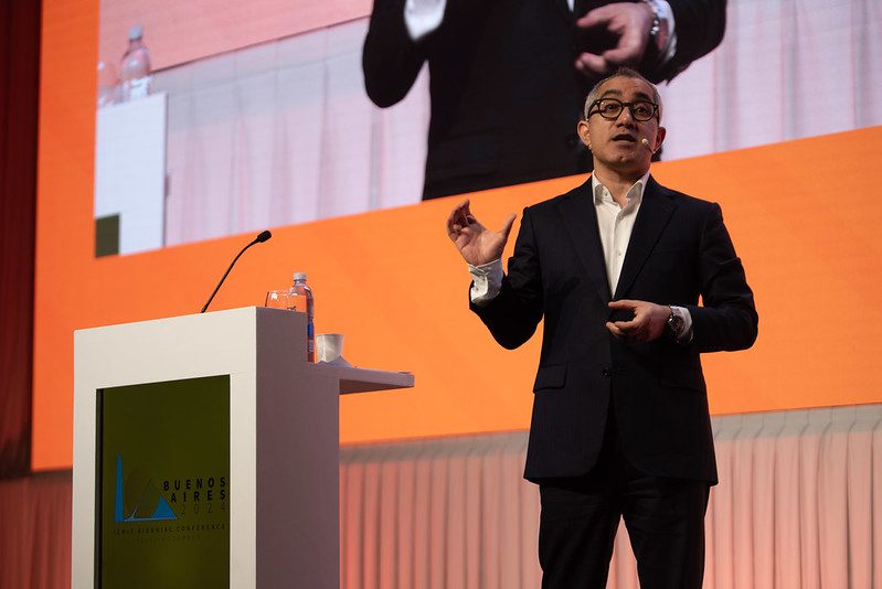 A man in a suit speaks on stage next to a podium with a “Buenos Aires” sign; a large screen behind him displays his image as he gestures with one hand.