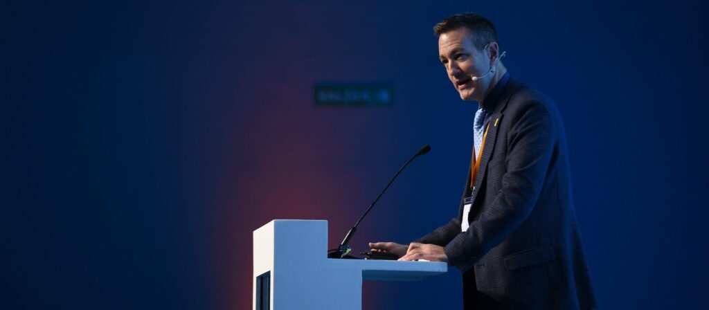 A man in a suit speaks at a podium with a microphone in a dimly lit room, against a blue background.