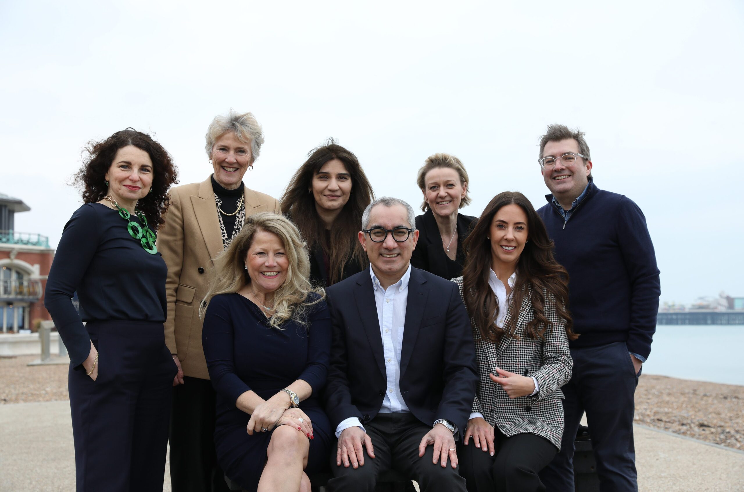 A group of eight professionally dressed adults pose and smile outdoors near a beach, with a pier and cloudy sky in the background. Four people are standing and four are seated in front.