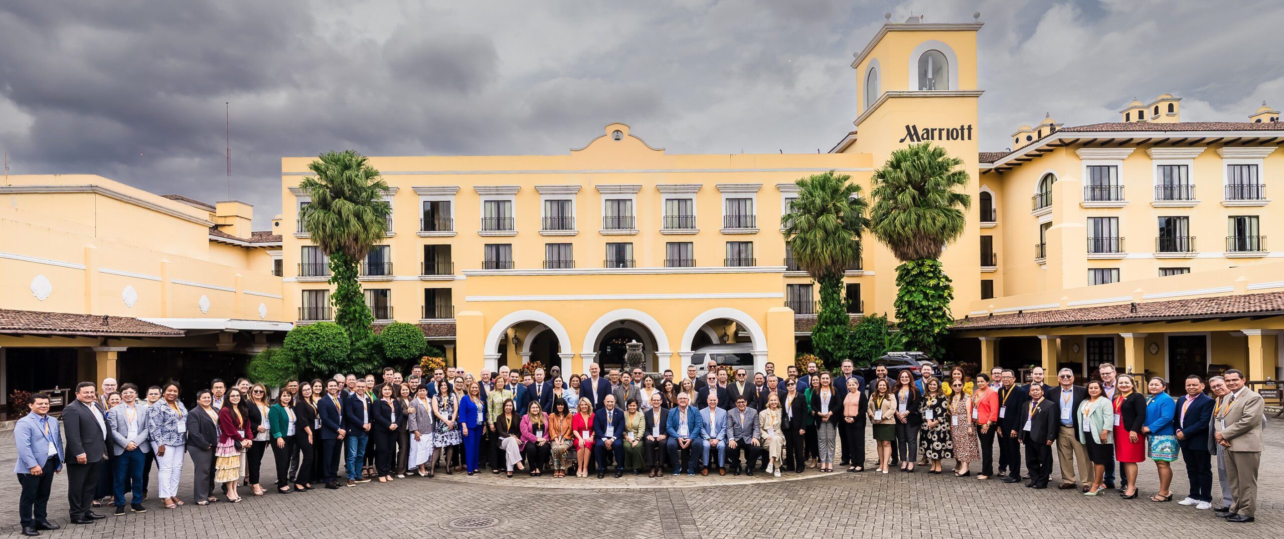 A large group of people in formal attire pose for a group photo in front of a yellow Marriott hotel building with arches, palm trees, and a cloudy sky overhead.
