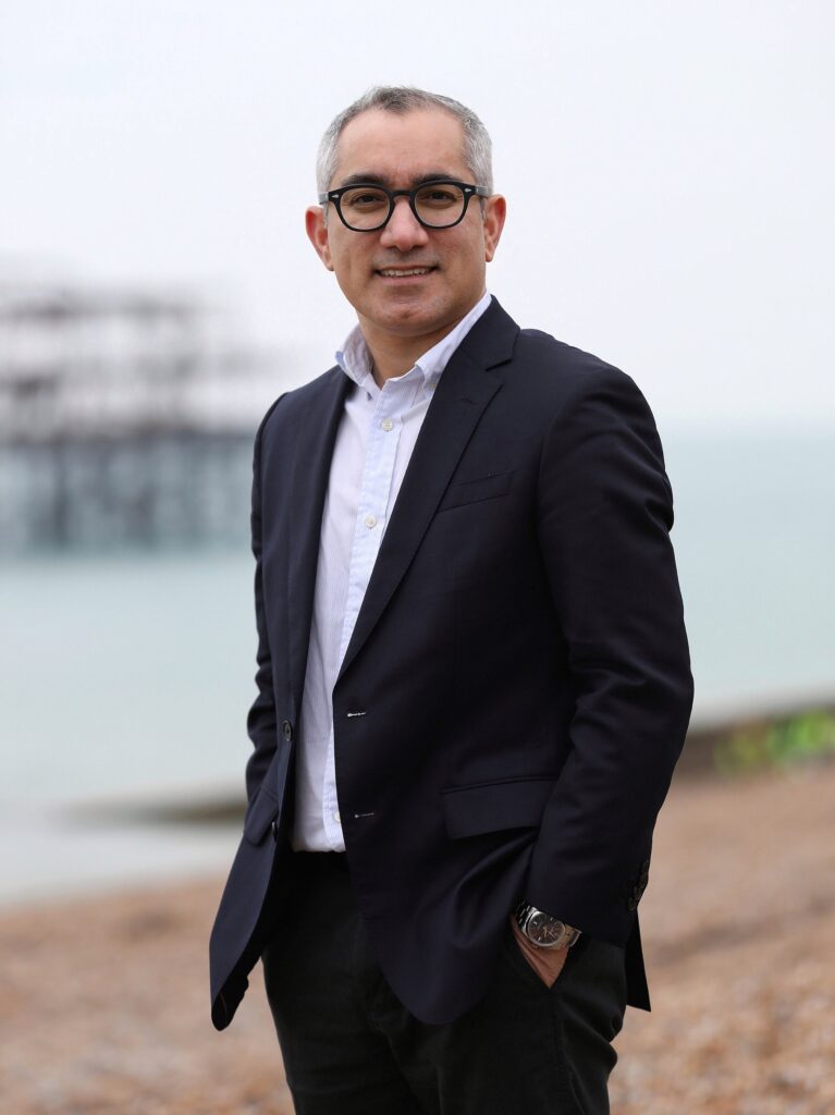 A man wearing glasses, a white shirt, and a dark blazer stands on a pebbled beach with a calm sea and an old pier in the background, smiling with his hands in his pockets.