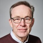 A middle-aged man with short brown hair, glasses, and a light complexion, wearing a dark jumper over a collared shirt, looks directly at the camera against a plain grey background.