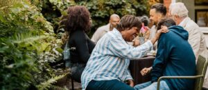 A group of people sit outside at a garden table, laughing and enjoying each other's company. One woman in a striped shirt leans forward, smiling and touching the shoulder of a person in a blue hoodie.
