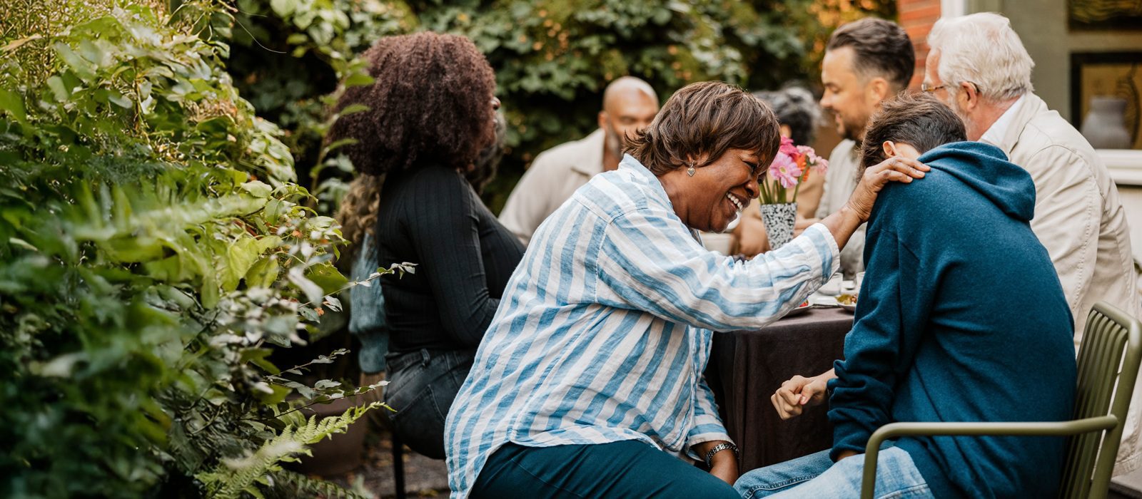 A group of people sit outside at a garden table, laughing and enjoying each other's company. One woman in a striped shirt leans forward, smiling and touching the shoulder of a person in a blue hoodie.