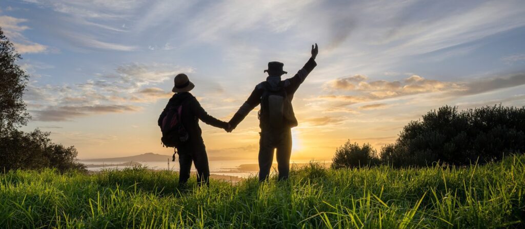 Two people holding hands stand in a grassy field at sunset, with one waving. Both wear hats and rucksacks, facing the horizon with a view of distant water and silhouetted trees under a colourful sky.