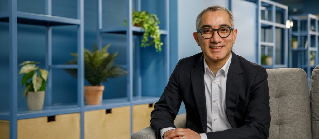 A man in a suit and glasses sits on a gray chair, smiling, with blue shelving and potted plants in the background. The setting appears to be a modern office or lounge area.