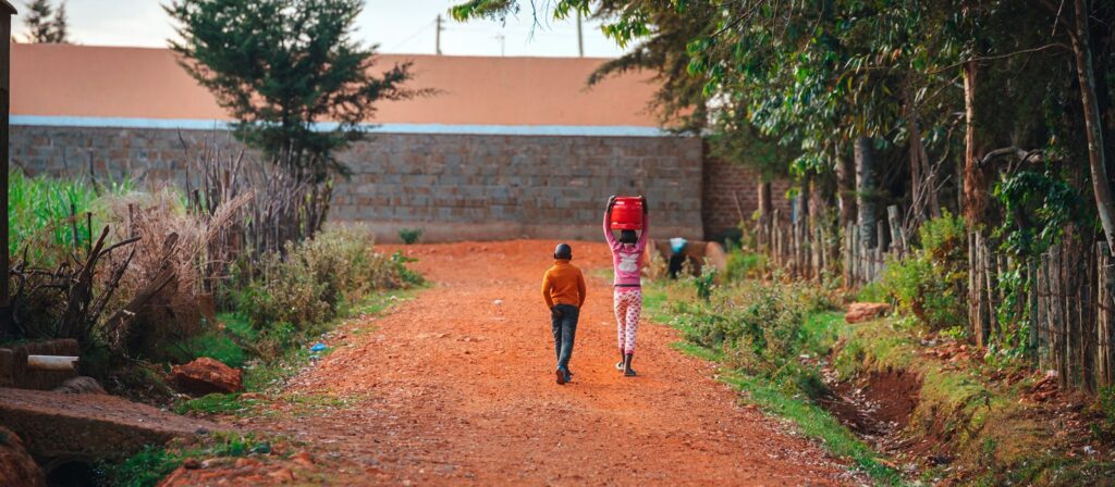 Two children walk down a dirt road. One child wears an orange hoodie, and the other, in pink clothes, carries a large red container on their head. Greenery lines the path, and a stone wall is in the background.