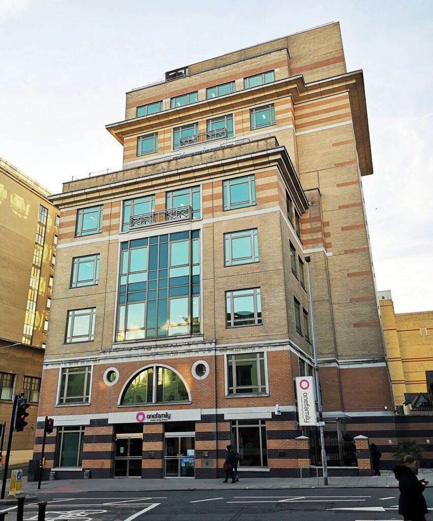 A multi-story modern office building with large windows and a mix of tan and red brick exterior. The onefamily sign is visible above the entrance. The building is located at a street corner in an urban area.