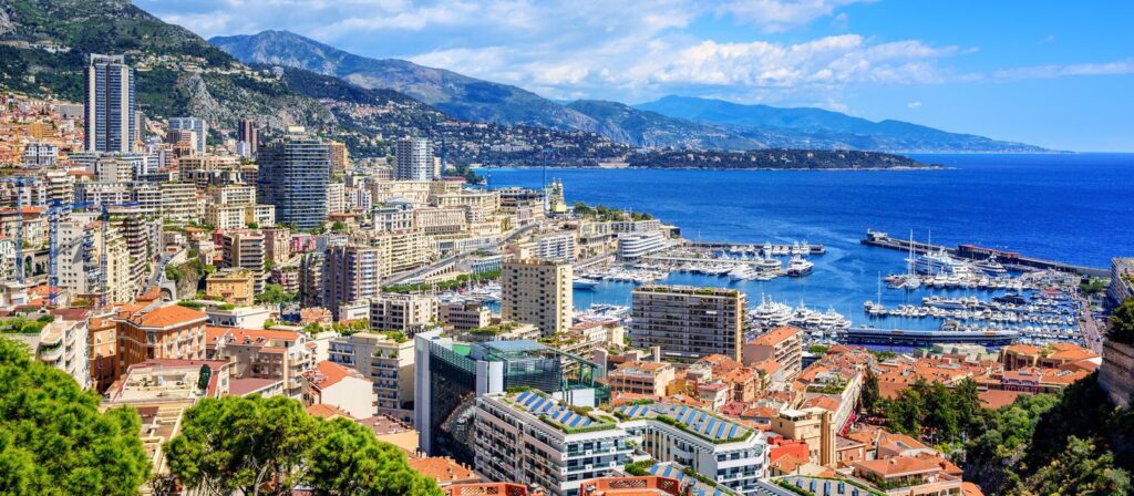 A panoramic view of Monte Carlo, Monaco, showing tall buildings, luxury yachts in the marina, and the deep blue Mediterranean Sea, with mountains in the background under a partly cloudy sky.