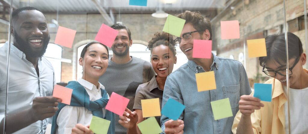 A group of six diverse people smiling and standing behind a glass wall covered with colourful sticky notes, appearing to brainstorm or collaborate in a modern office setting.