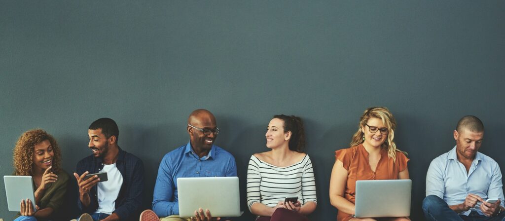 Six people sit in a row against a grey wall, smiling and interacting with each other. Some are using laptops, tablets, or mobile phones, while others chat. They appear relaxed and engaged, suggesting a collaborative or work setting.