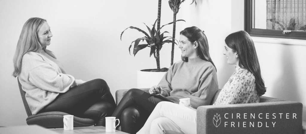 Three women sit together in a casual office setting, engaged in conversation with coffee cups in hand. A plant and a window are in the background. The Cirencester Friendly logo is visible in the bottom right corner.