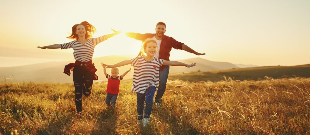 A happy family of four, with arms outstretched, runs through a sunny field at sunset. The parents and two children smile as they play together in the tall grass, mountains visible in the background.