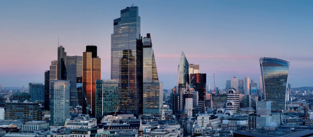 A panoramic view of London’s modern skyline at sunset, featuring tall glass skyscrapers, including The Gherkin and The Walkie-Talkie, with older historic buildings in the foreground.