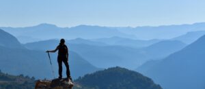 A person stands on a rocky ledge with a walking stick, overlooking layers of blue mountains under a clear sky, enjoying the scenic view.