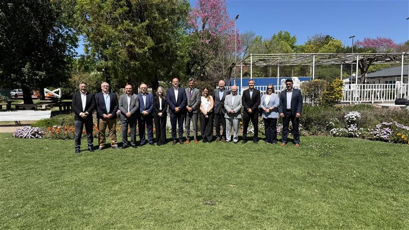 A group of fifteen people, mostly in suits, stand in a row on a green lawn with flowers, trees, and a white structure in the background, under a clear blue sky.