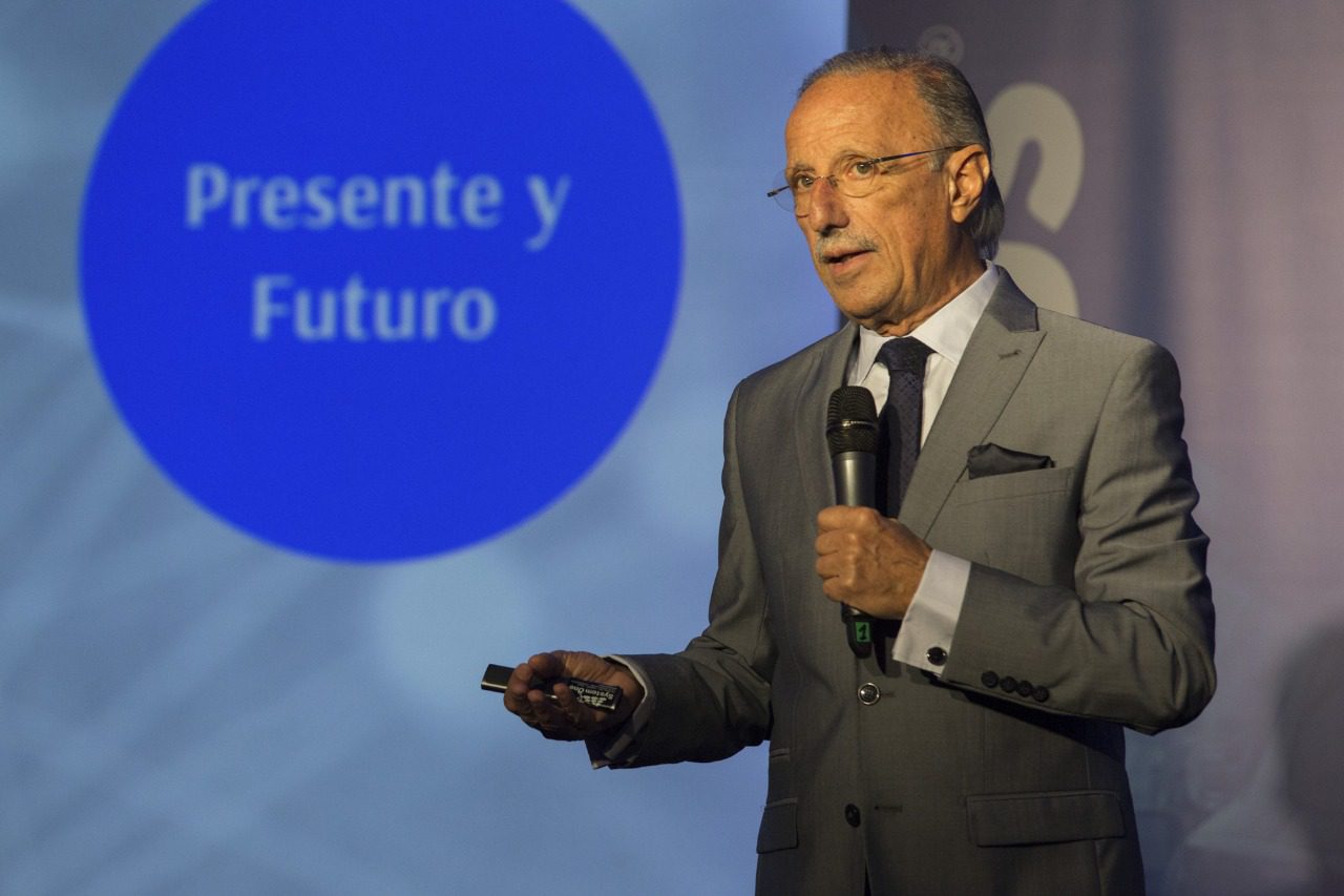 A man in a grey suit and tie holds a microphone and remote, speaking in front of a screen displaying a blue circle with the Spanish text “Presente y Futuro”.
