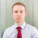 A man with short light brown hair wearing a light blue shirt and red patterned tie sits in front of a blurred office background, looking directly at the camera with a neutral expression.