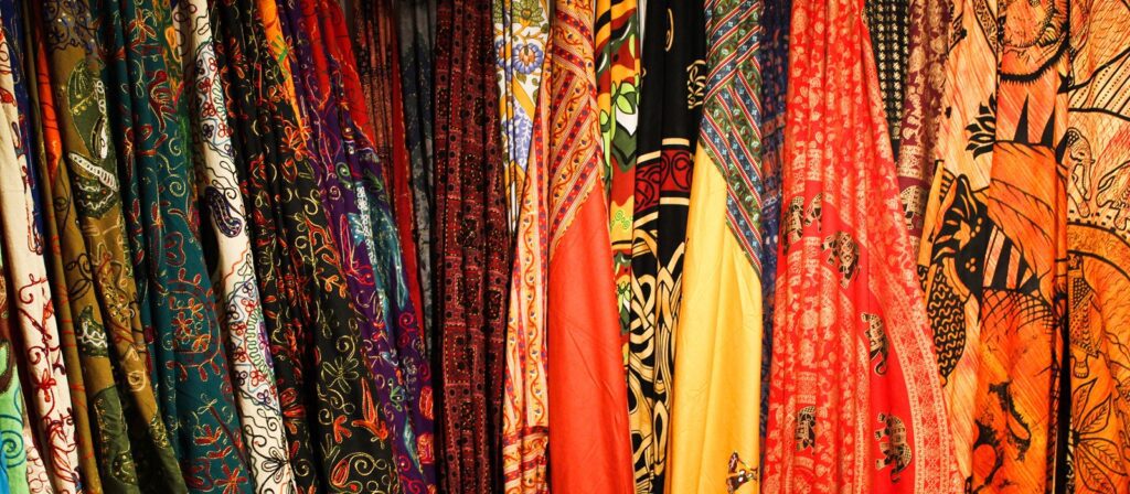 A colourful display of patterned fabrics hanging vertically, featuring intricate designs and a variety of vibrant colours including red, orange, yellow, green, blue, and black.