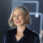 A smiling older woman with short grey hair, red lipstick, and gold jewellery is wearing a dark silk blouse. She poses in front of a blurred, modern background.