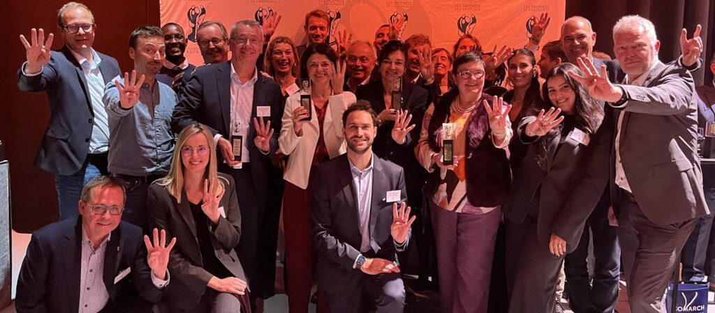 A group of smiling people in business attire pose together, many holding up four fingers. They stand and crouch in front of a red backdrop with logos, suggesting a celebratory or award event.