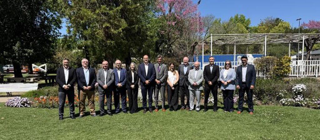 A group of fifteen people, dressed in business attire, stand in a row outdoors on a grassy area with trees, flowers, and structures in the background on a sunny day.