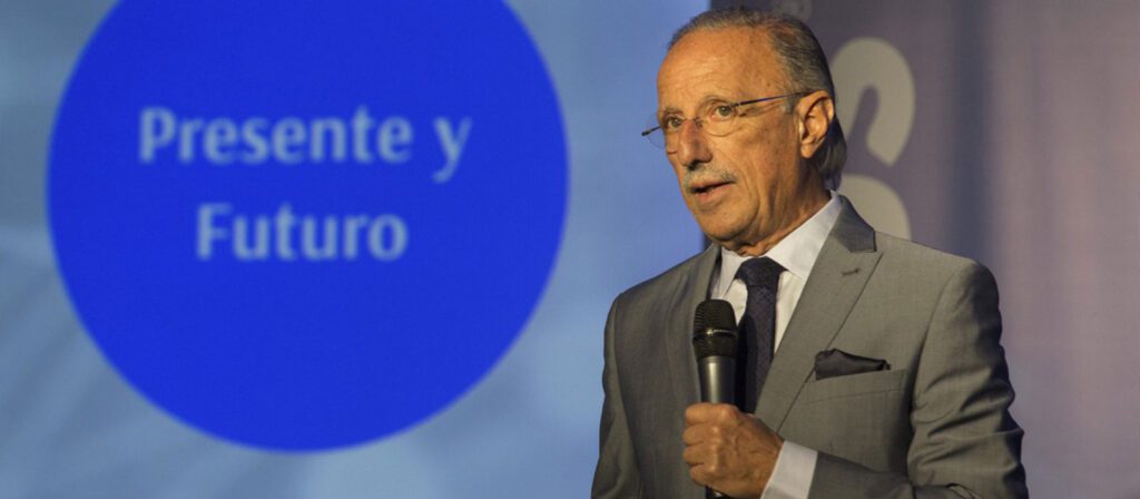 A man in a grey suit and tie speaks into a microphone. Behind him, a blue circle displays the Spanish words Presente y Futuro on a screen.