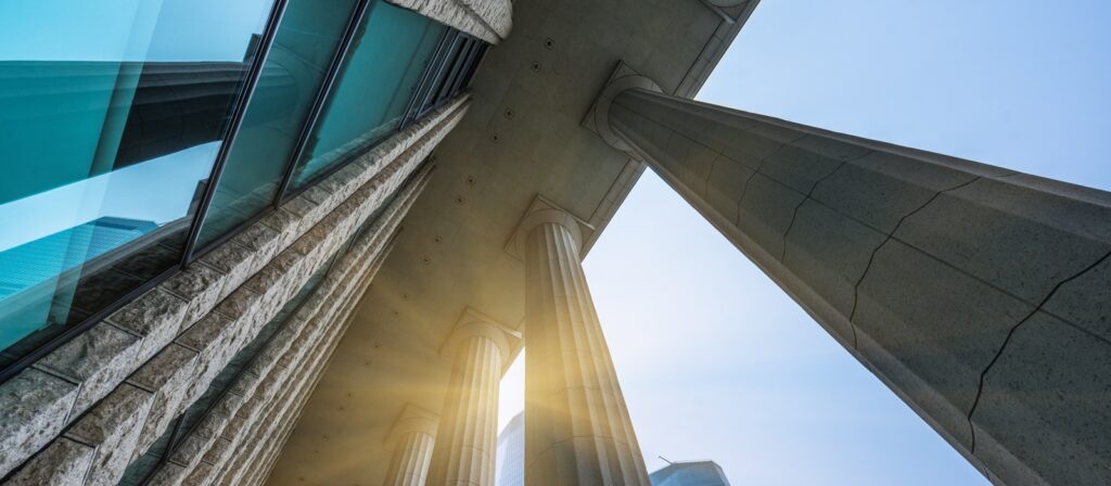 Low-angle view of a modern building with tall stone columns and reflective glass windows, sunlight streaming between the columns, and a clear blue sky in the background.
